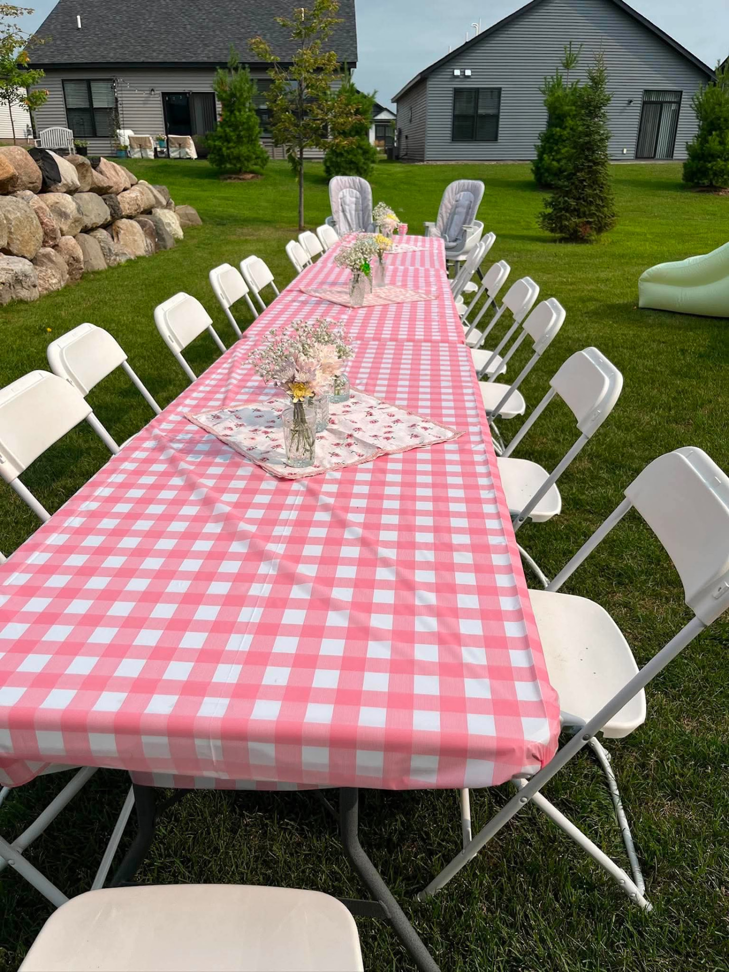 Long banquet table with pink gingham linen and white folding chairs in a backyard